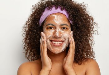 Positive pleasant looking dark haired Afro woman with bare shoulders massages face, washes with soap
